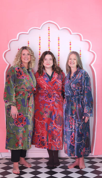 Three women wearing colorful patterned robes standing in front of a decorative pink wall.